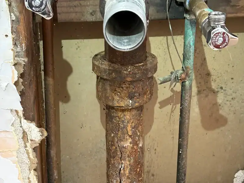 Corroded cast iron drain riser inside an opened bathroom wall, with a crack running down the pipe